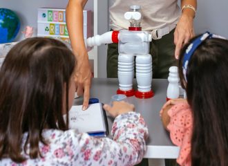 Female teacher in ecology classroom explaining to children how make recycled toy robot with plastic packages